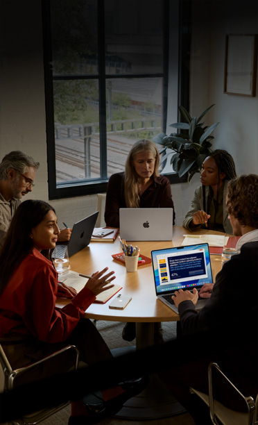 Cinq personnes assises autour d’une table de conférence travaillent ensemble sur des MacBook, leurs iPhone face vers le bas