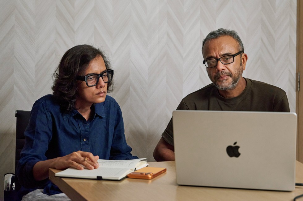 Dhritisree Sarkar and a colleague sit at a table, looking at a MacBook Pro. iPhone 17 Pro Max sits on the table beside them.