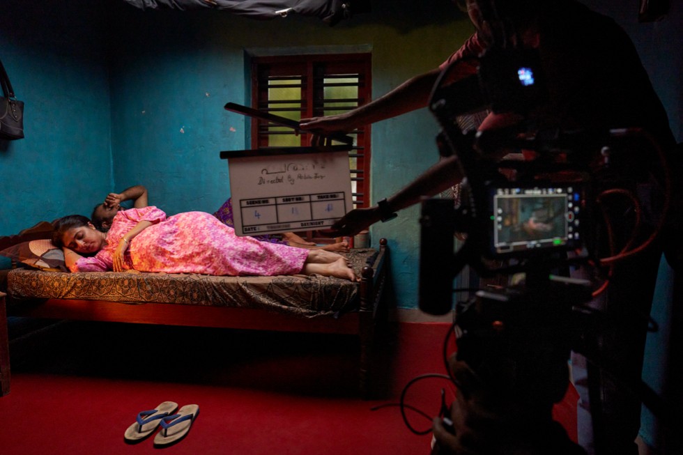 A film set showing a person lying on a bed in a dimly lit room, with camera equipment visible.