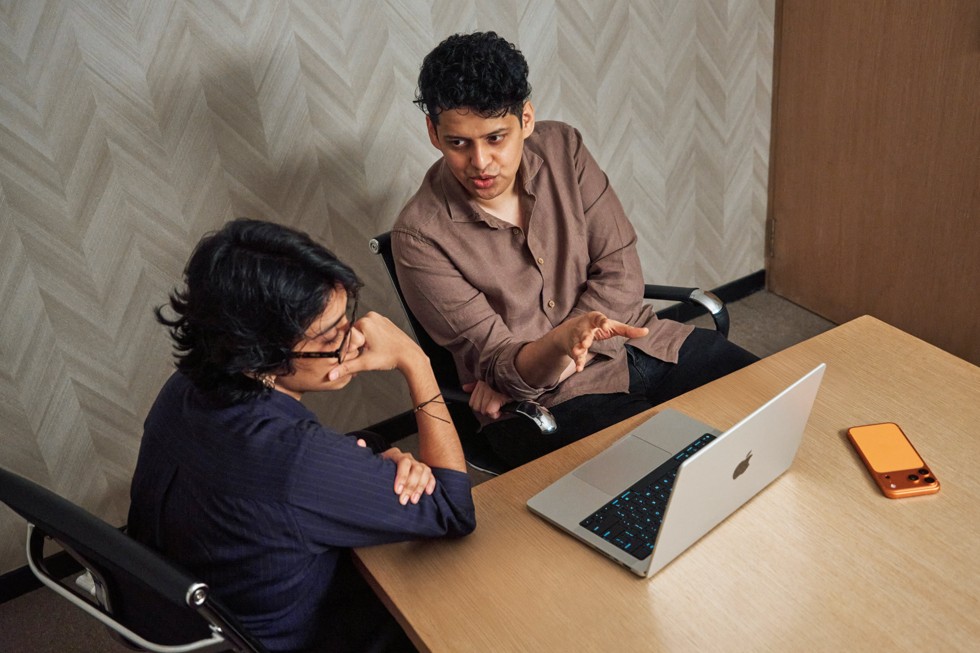 Shreela Agarwal sits at a table with mentor Chaitanya Tamhane, looking at a MacBook Pro and having a discussion. iPhone 17 Pro Max sits on the table beside them.