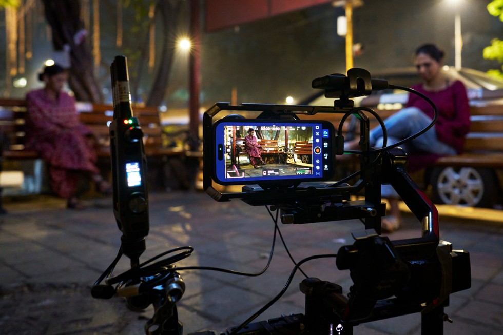 iPhone 17 Pro Max on a camera rig is mounted on a tripod, filming a scene at night with a person sitting on a bench in the background.