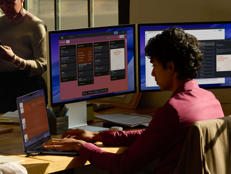 Un equipo de empleados sentados alrededor de una mesa en una oficina. Una mujer sentada en su escritorio trabaja en una MacBook con varios monitores. Sus colegas están reunidos conversando.