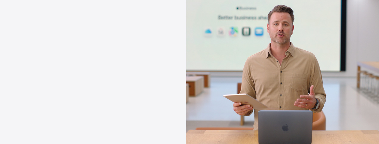 Un presentador habla con un iPad en la mano y un MacBook delante sobre una mesa, mientras que en una pantalla grande se muestran los iconos de varias apps de trabajo.