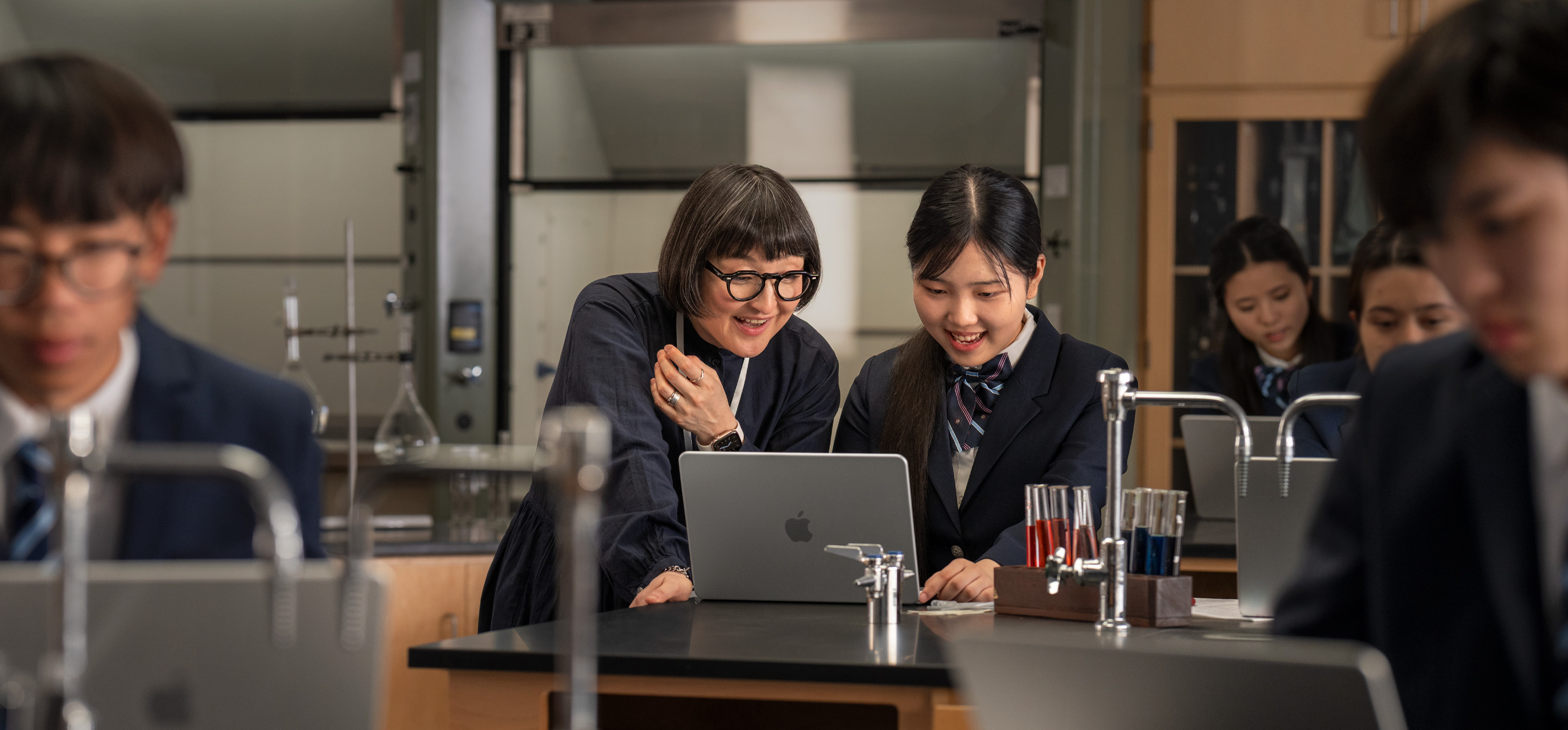 試験管や実験器具に囲まれた理科室で、机の上に開かれたMacBookを前に対話している生徒と教師