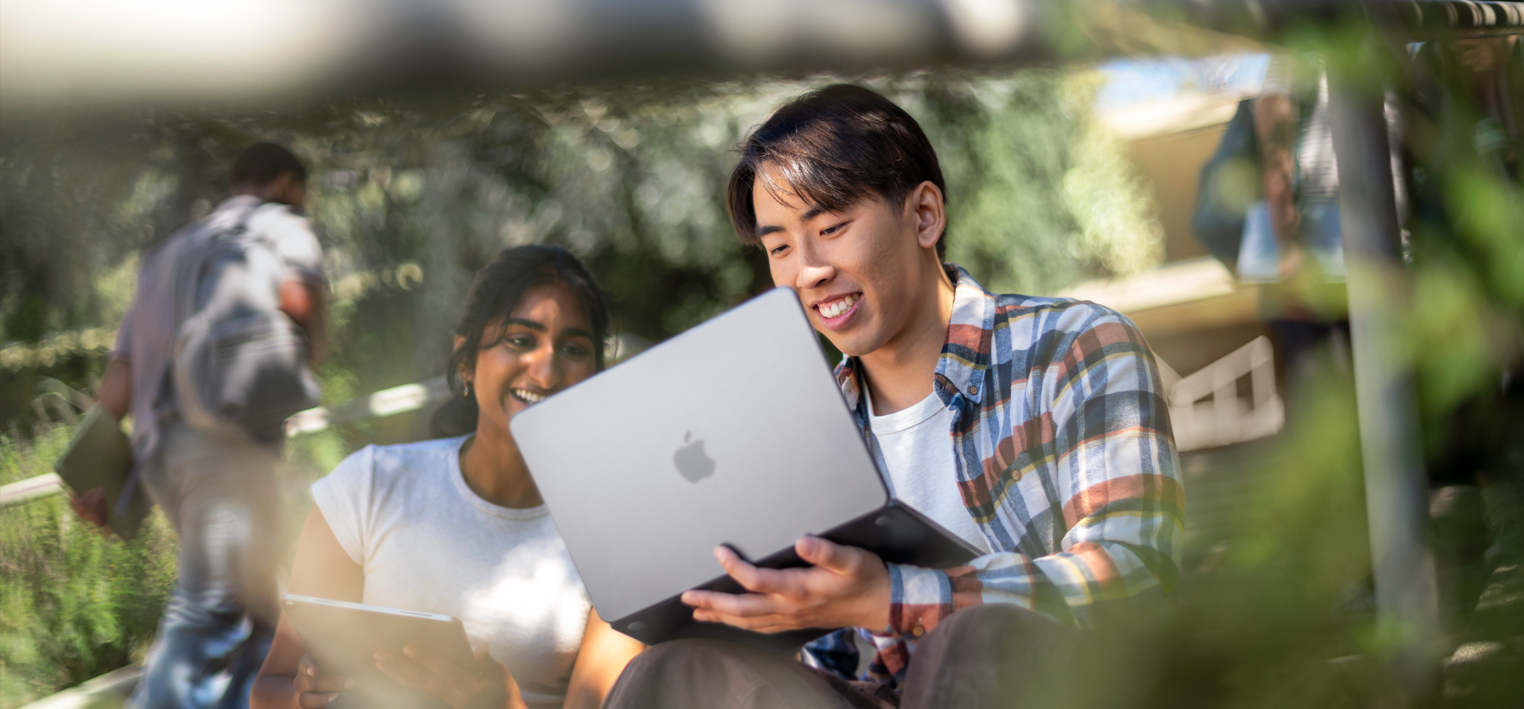 屋外でMacBookを使いながら、笑顔で課題の共同作業に取り組んでいる2人の学生。1人は膝の上にMacBookを置いている