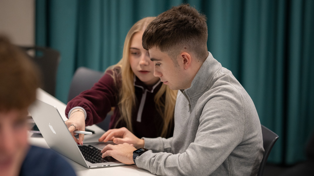 Klasgenoten werken samen op een MacBook, de een wijst naar het scherm terwijl de ander typt