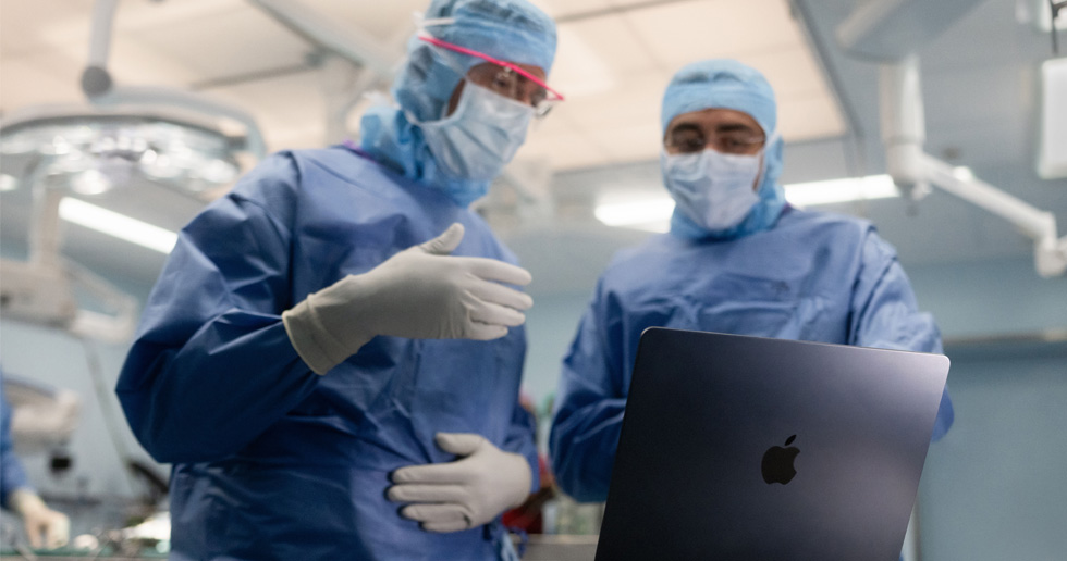 Zwei Chirurgen im Operationssaal schauen auf ein MacBook, wobei einer eine Geste mit der Hand macht, während der andere beobachtet