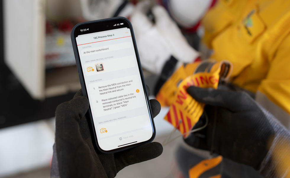 Unas manos sostienen un iPhone que muestra instrucciones para realizar una prueba de cuadro eléctrico, con las palabras 13C Proceso Paso 3 en la pantalla