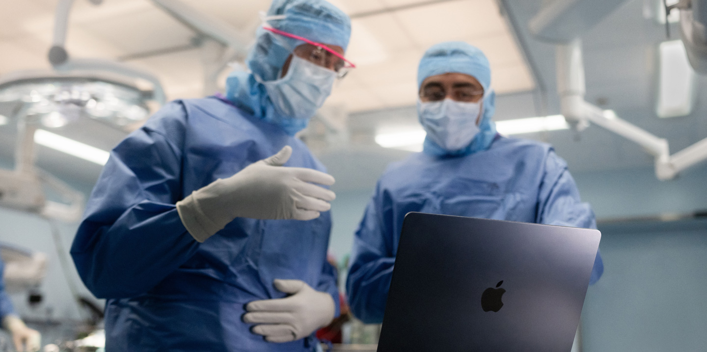 To kirurger i et operasjonsrom ser på en MacBook, og den ene gestikulerer mens den andre ser på