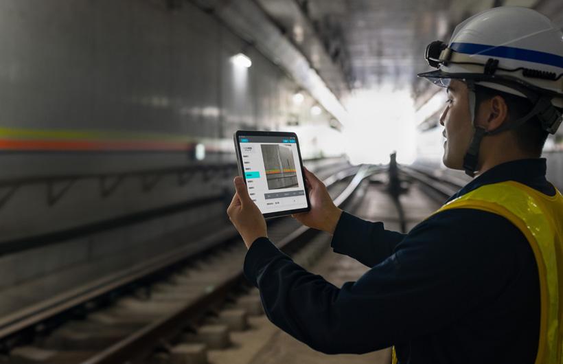 En underhållstekniker i en av Tokyo Metros tunnlar håller upp en iPad för att ta en bild av spåren medan en kollega tittar på.