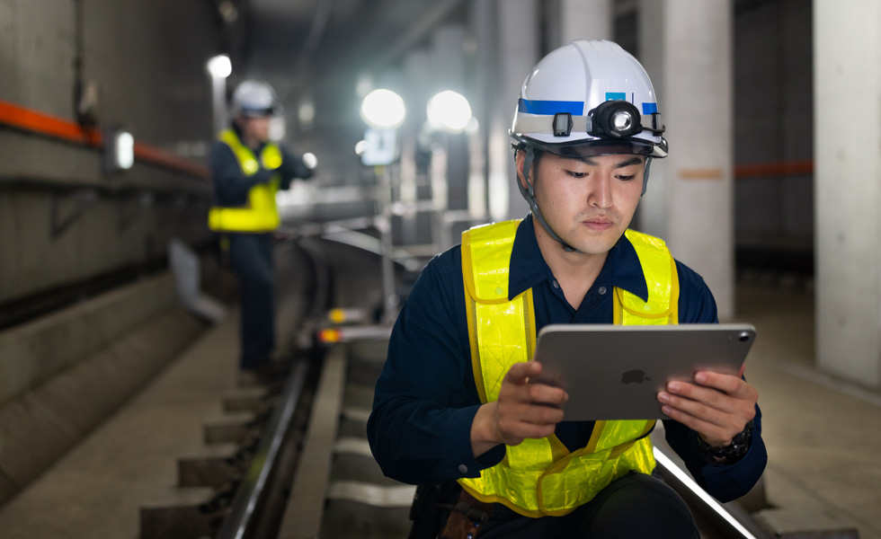A worker wearing a safety vest and hard hat looks at an iPad while performing maintenance in the tunnels of Tokyo Metro.